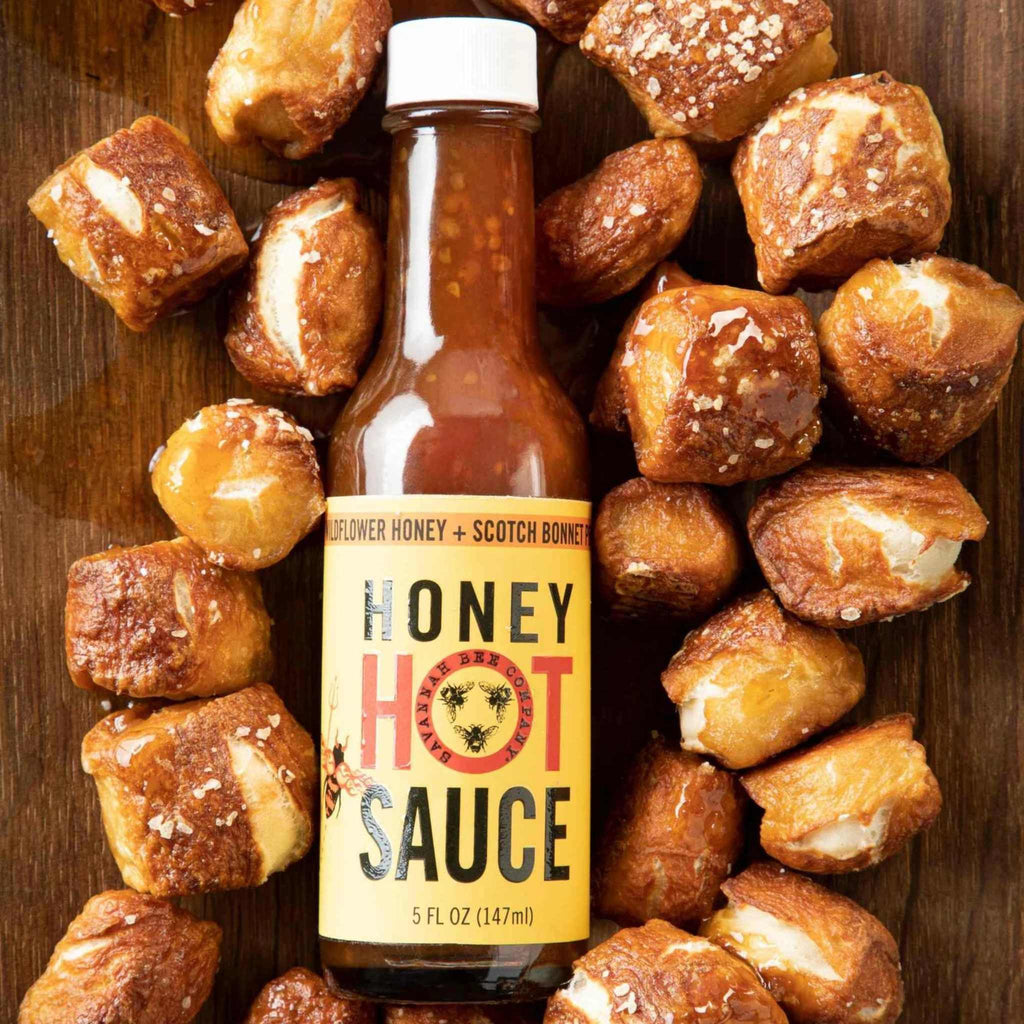 Honey Hot Sauce 5 oz. bottle from the Savannah Bee Company laying on a wood table surrounded by pretzel bites.